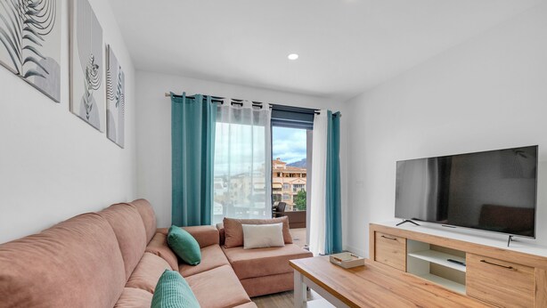 A modern living room with a sofa, a TV, a wooden table, and a view of buildings through the window. - LXR Meninas 2-Bedroom Apartment Near the Sea (Fuengirola)