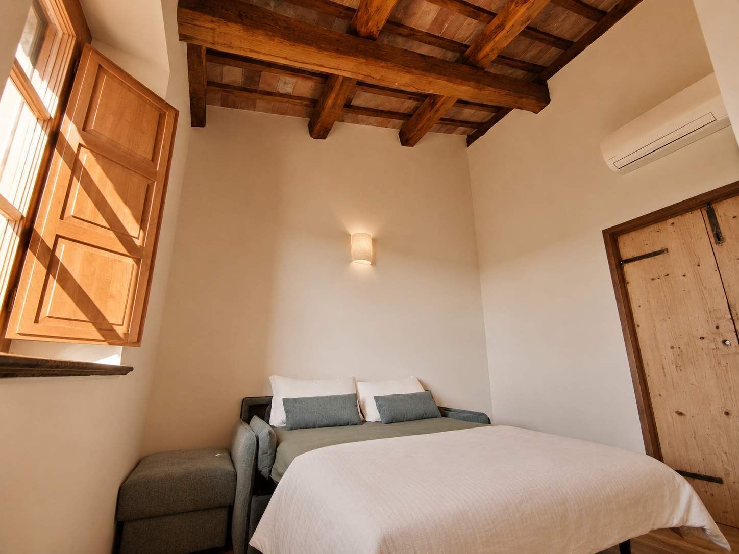 Un dormitorio con cama, mesita de noche, ventana con contraventanas de madera, techo con vigas a la vista y unidad de aire acondicionado instalada en la pared.