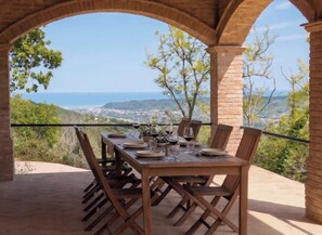 An outdoor dining area with a table set for four, overlooking a scenic view of the sea and hills.
