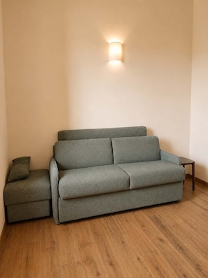 A grey sofa with a matching ottoman and a small table, against a plain wall with a wall-mounted light fixture. - Villa Santa Maria dell'Olmo (MORESCO)