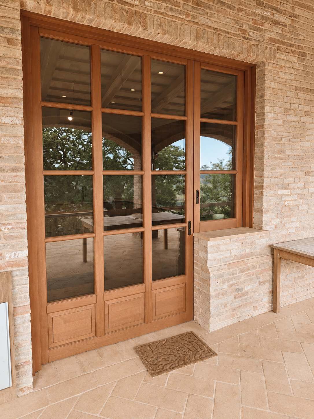 A large wooden door with glass panels, set in a brick wall with a stone bench and a doormat in front.