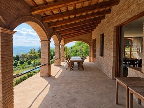Eine überdachte Terrasse im Freien mit Tisch und Stühlen, Backsteinmauern und Blick auf Grünflächen und Berge.