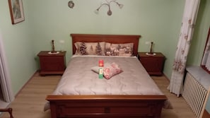 Quarto com cama, mesas de cabeceira, radiador e janela com cortinas.