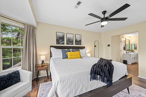 Ein Schlafzimmer mit Bett, Deckenventilator, Nachttisch, Fenster mit Vorhängen und angrenzendem Badezimmer.