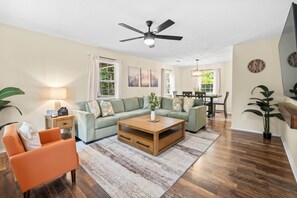 A living room with a sofa, coffee table, and orange armchair, a dining area with a table and chairs, and a ceiling fan. - Mountain City Hideaway - Downtown Asheville Retreat with Game Room, Deck + Grill (Asheville)