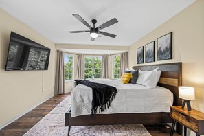 Ein Schlafzimmer mit Bett, Deckenventilator, Wandfernseher und einem fenster mit Vorhängen.