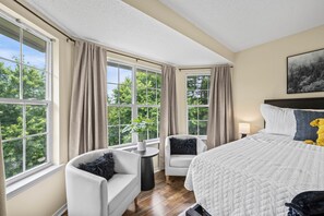 Ein Schlafzimmer mit einem großen Bett, zwei Stühlen, einem Nachttisch, einer Lampe und einem Fenster mit Vorh ängen.
