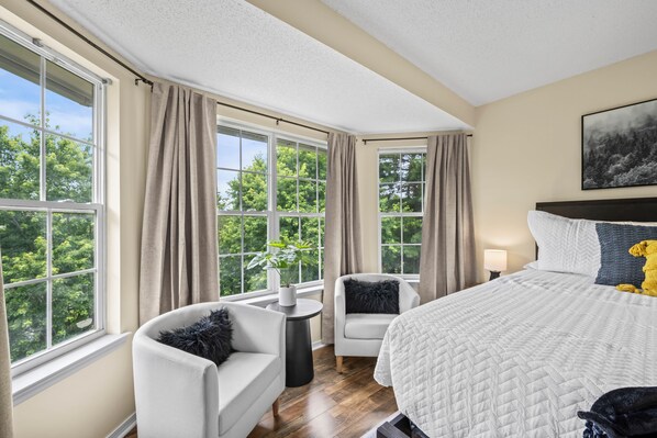 A bedroom with a large bed, two chairs, a bedside table, a lamp, and a window with curtains.