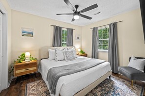 Quarto com cama, mesas de cabeceira, uma cadeira, ventilador de teto e janela com cortinas.