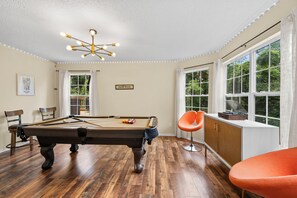 Uma mesa de bilhar em um quarto com luminária no teto, uma cadeira e uma janela com cortinas.