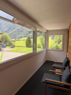 Ein Zimmer mit großen Fenstern, das einen Blick auf eine grüne Landschaft und Berge bietet. Es gibt zwei Holzstühle mit Kissen.