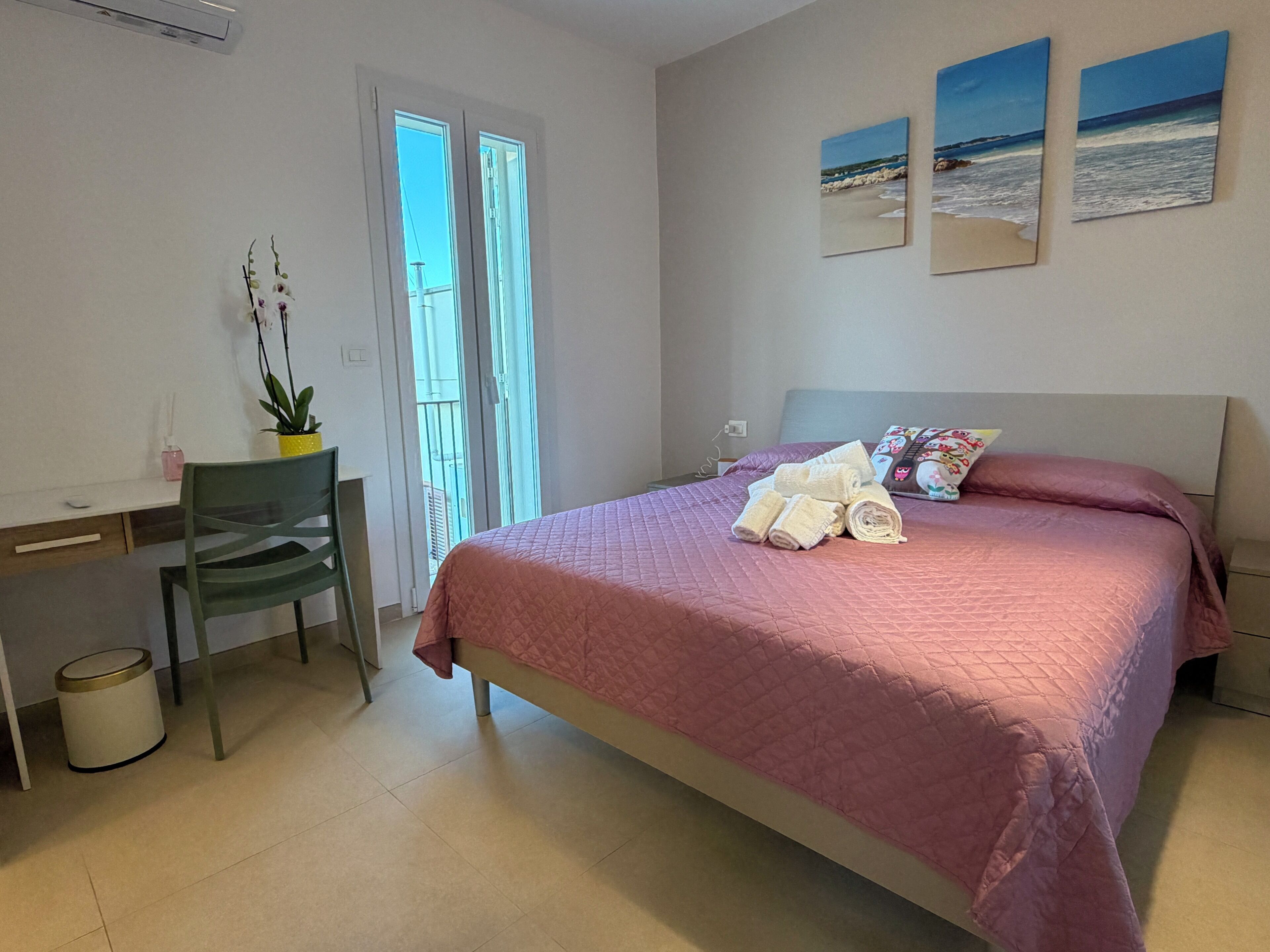 Een slaapkamer met een bed, een bureau, een stoel, een potplant en twee schilderijen aan de muur.