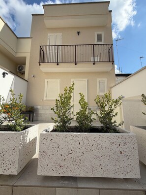 Ein zweistöckiges Gebäude mit einem Balkon, weißen Fensterläden und einem Garten mit Pflanzkübeln.