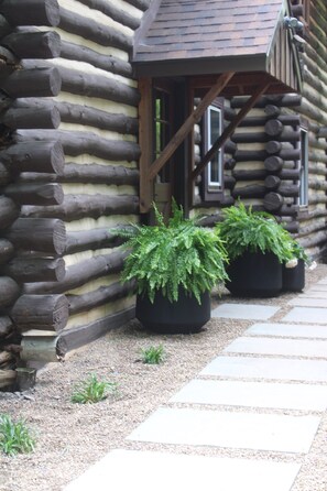 Une cabane en rondins avec un escalier en bois menant à une porte, deux fougères en pot et un chemin de gravier.