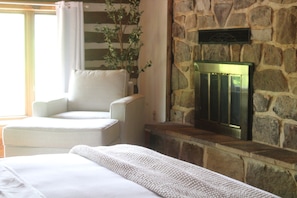 Une pièce avec une cheminée en pierre, un fauteuil blanc et un lit recouvert d’une couverture blanche.