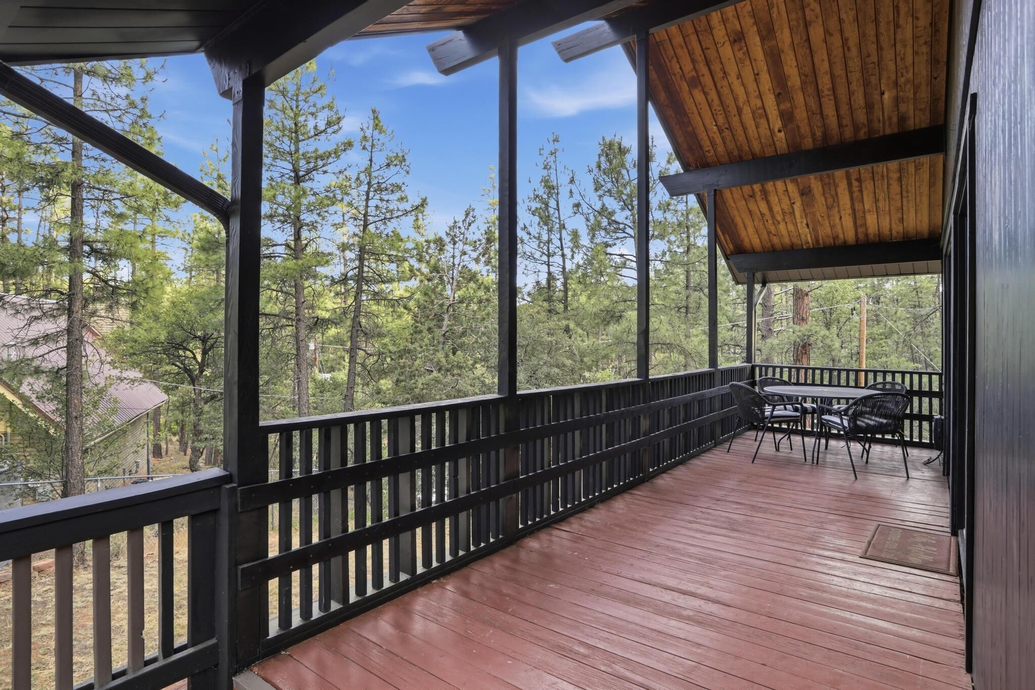 A covered outdoor deck with a wooden floor, black railings, and a view of tall trees.