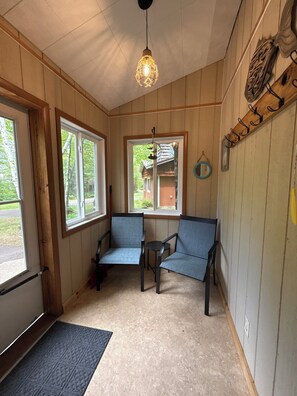 A room with wooden walls, a ceiling light, two chairs, a small table, and a window with a view of a house. - Superior View at Miller's Cottages (Ontonagon)