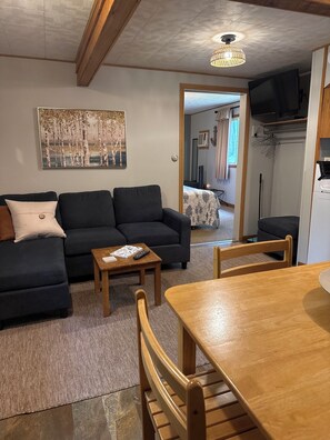 A living room with a sofa, coffee table, and dining table. There is a painting on the wall and a ceiling light fixture. - Superior View at Miller's Cottages (Ontonagon)