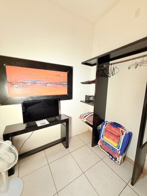 Ein Zimmer mit einem an einer schwarzen Halterung befestigten Fernseher, einem weißen Ventilator, einem gefliesten Boden und einem Stauraum mit Kleidung sowie einem farbenfrohen Stuhl.
