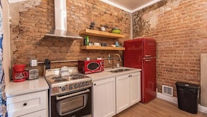 Une cuisine de taille réduite avec un mur en briques, un réfrigérateur rouge, un four à micro-ondes, une cuisinière et des armoires blanches.