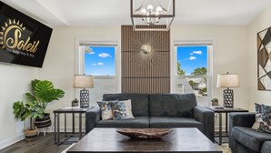 A modern living room with a grey sofa, two side tables with lamps, a wooden coffee table, and a large wall-mounted TV displaying 'Soleil - The Better Way To Stay'. - Raven's Nest - Mins to Beach Heated Pool & Pet Friendly (Panama City Beach)