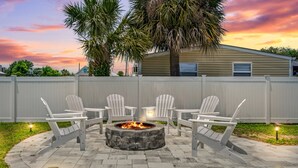 A patio with a stone fire pit surrounded by white Adirondack chairs, a white fence, and a house in the background. - Raven's Nest - Mins to Beach Heated Pool & Pet Friendly (Panama City Beach)