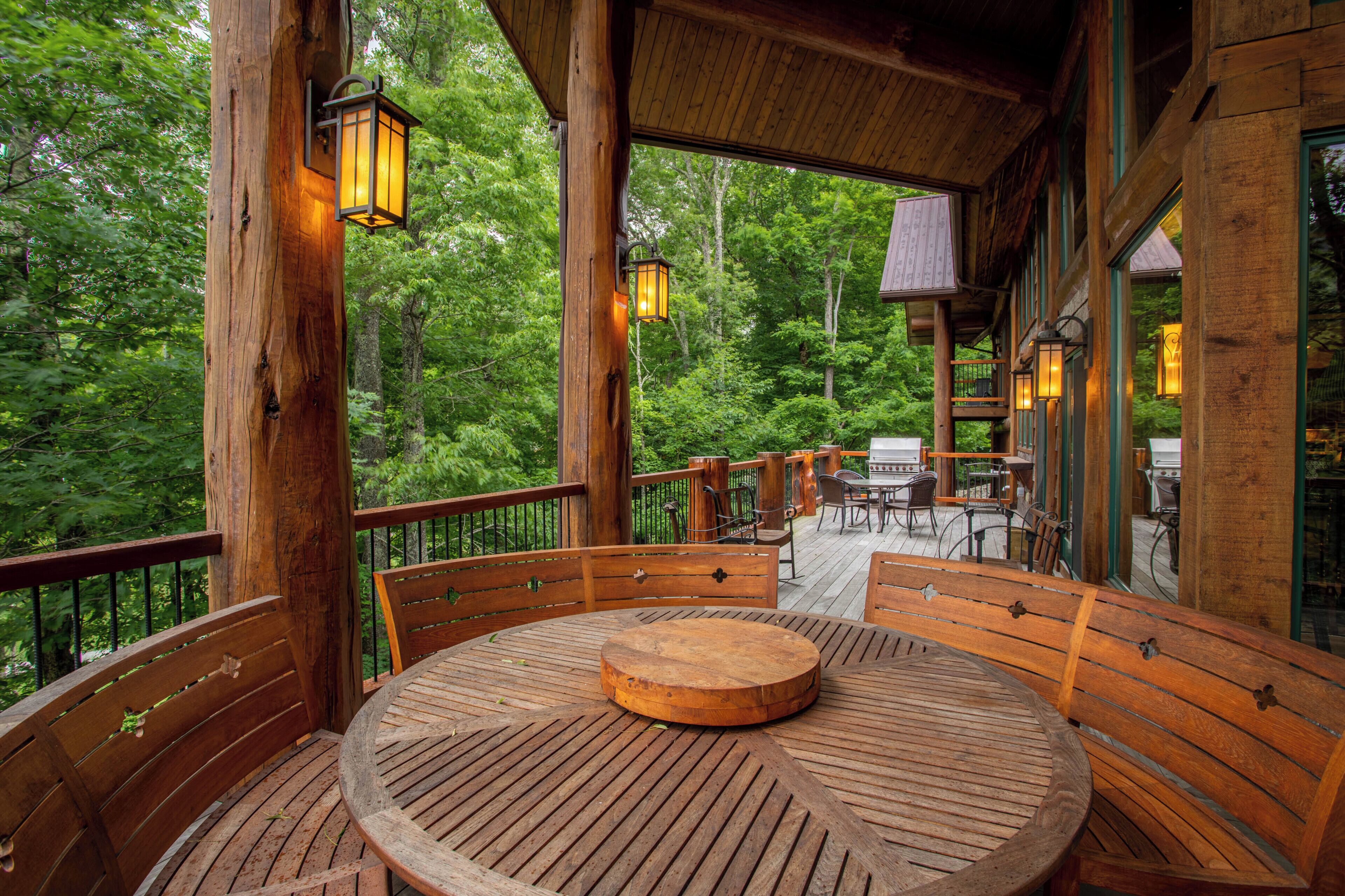 A wooden deck with a round table and benches, surrounded by glass windows offering a view of a forest.