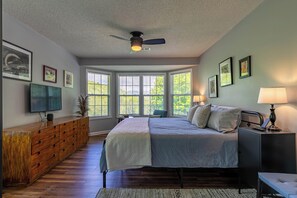 Un dormitorio con una cama, ventilador de techo, televisión, cómoda y ventanas amplias.