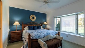Een slaapkamer met een bed, twee nachtkastjes, een plafondventilator, een raam met jaloezieën en uitzicht op een tennisbaan.