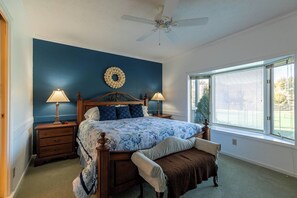 Un dormitorio con cama, dos mesitas de noche, ventilador de techo, ventana con persianas y vista a una cancha de tenis.