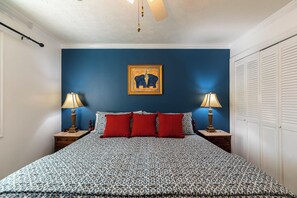 A bedroom with a patterned bedspread, two red pillows, a wall-mounted elephant painting, two bedside lamps, and a white closet with louvered doors.