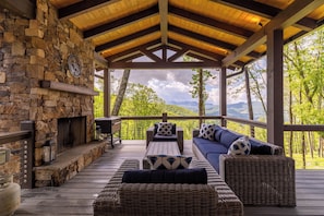 A covered outdoor seating area with stone walls, a fireplace, wicker furniture, and a view of green hills. - Howling at the Moon at Eagles Nest (Banner Elk)