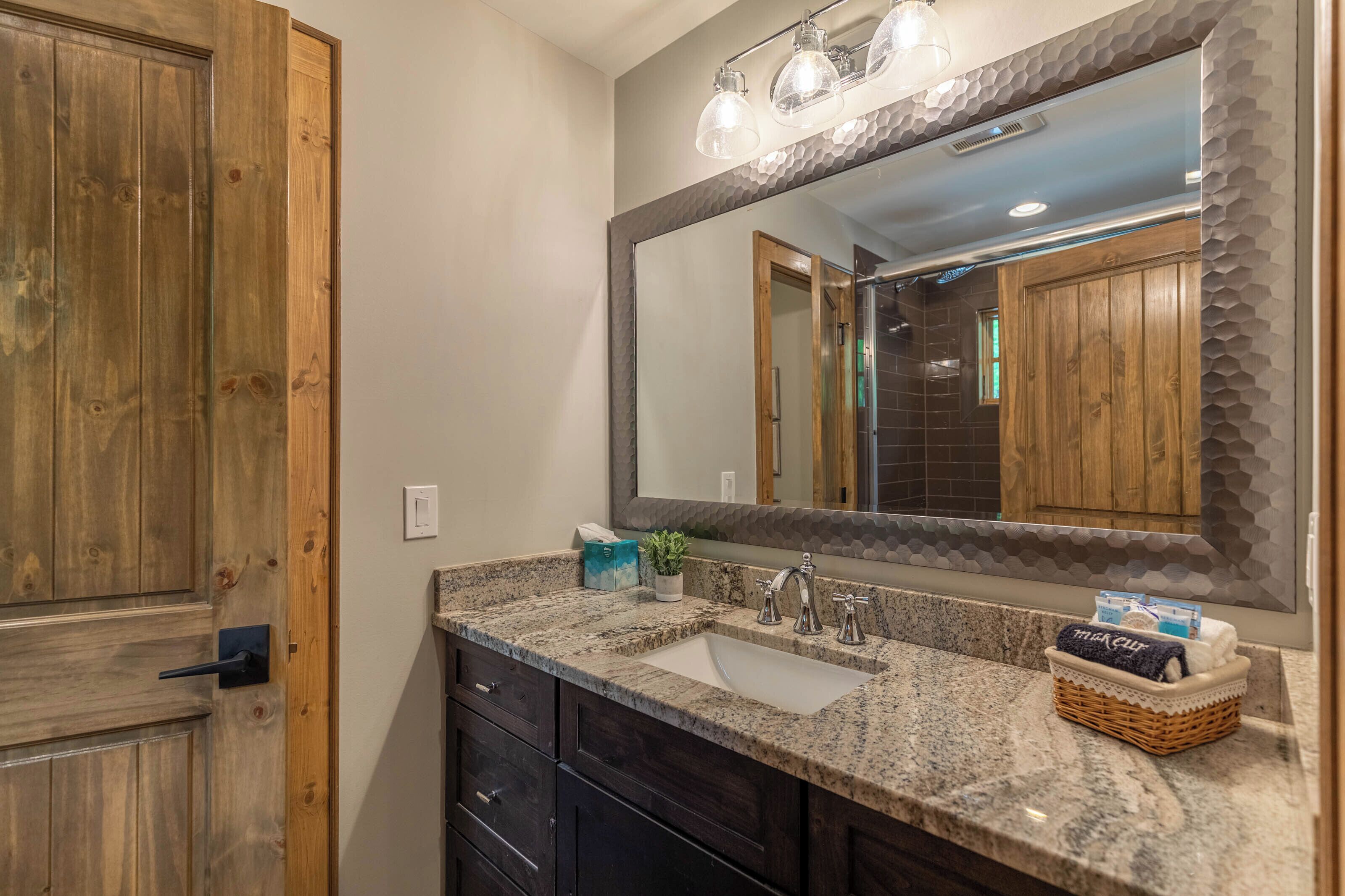 A bathroom with a wooden door, a granite countertop, a sink, a mirror, and a towel basket.