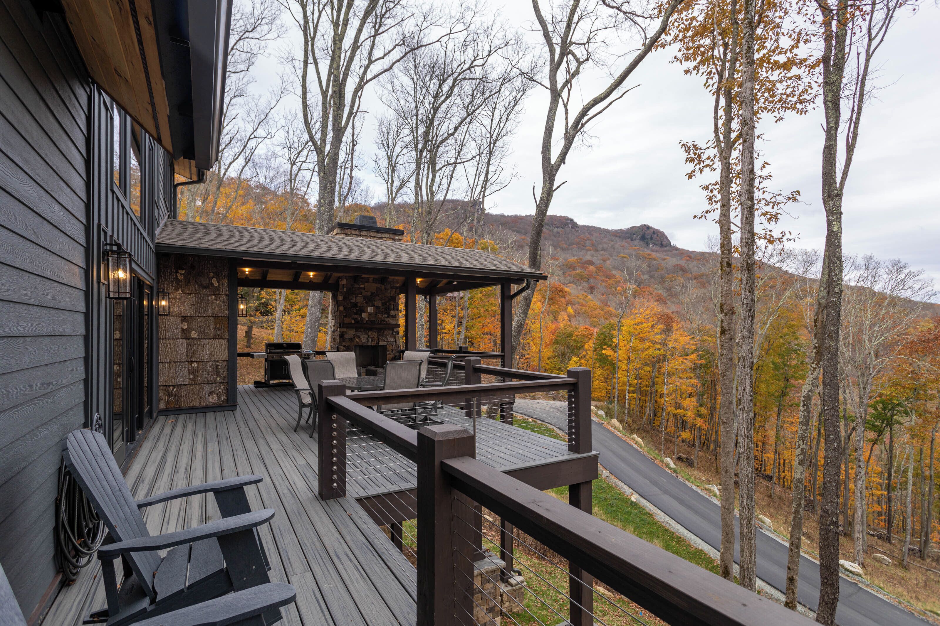 A wooden deck with outdoor seating and a stone fireplace, overlooking a forested area with autumn foliage.