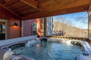 Una terraza de madera con un jacuzzi, barandillas de madera y vistas a árboles y una casa.