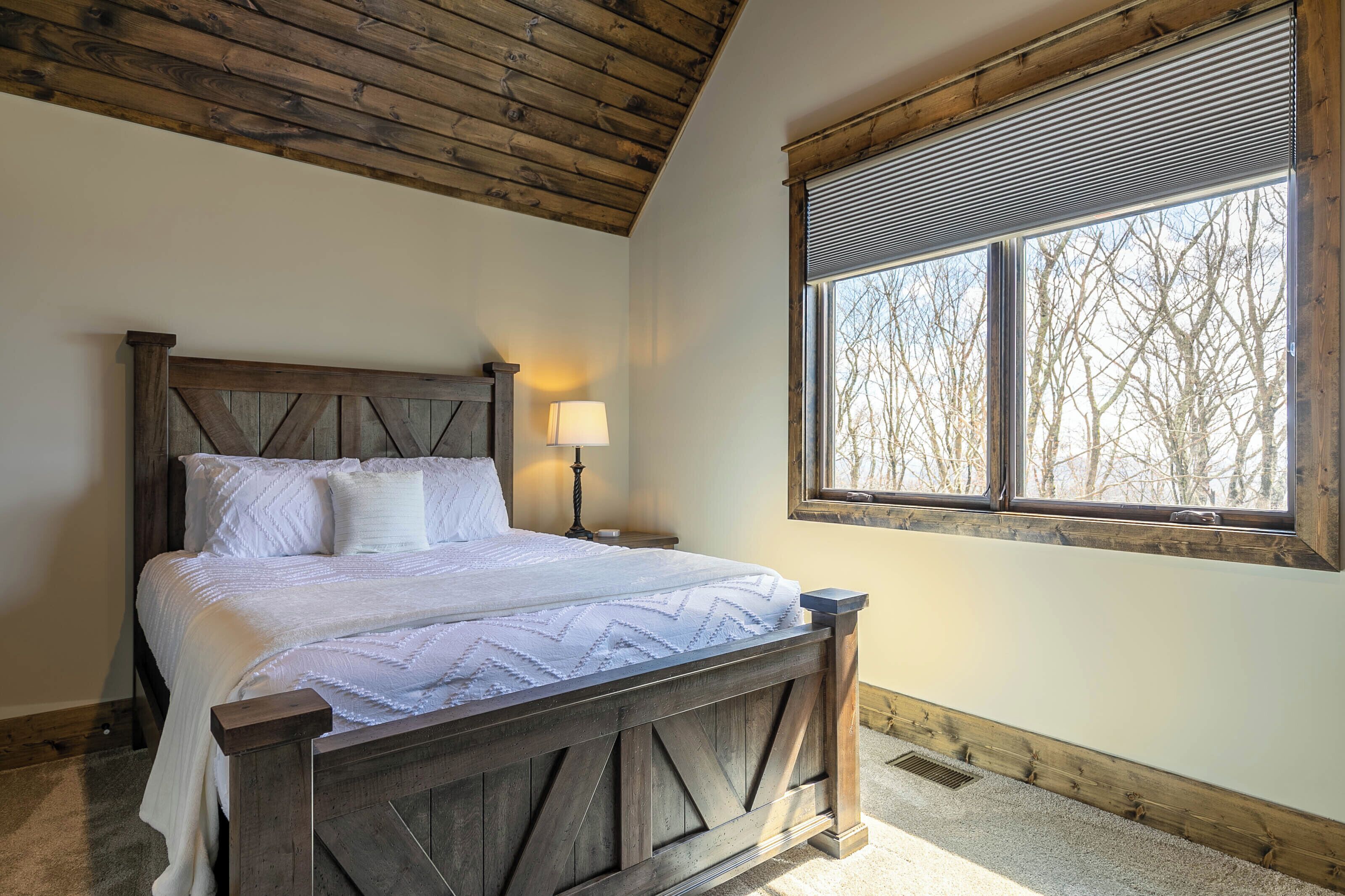 A bedroom with a wooden bed, a nightstand, a lamp, and a window with a view of trees.
