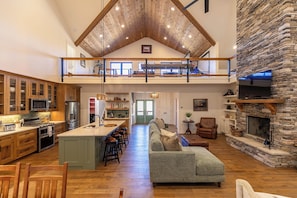A spacious loft-style living area with a high ceiling, a stone fireplace, a kitchen with modern appliances, and a cozy seating area.