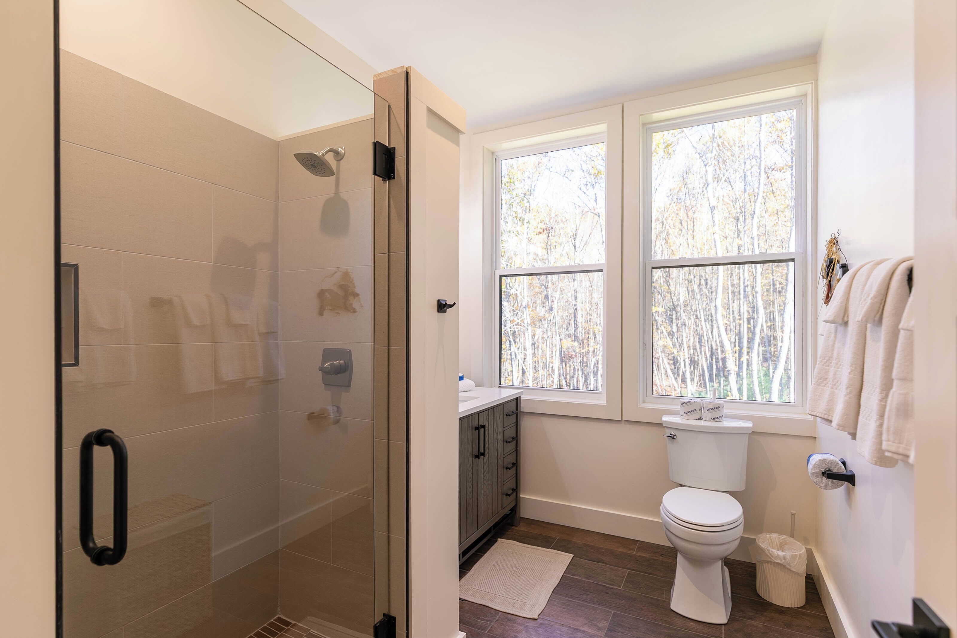 A bathroom with a glass shower enclosure, a toilet, a sink with a cabinet, and a window with a view of trees.
