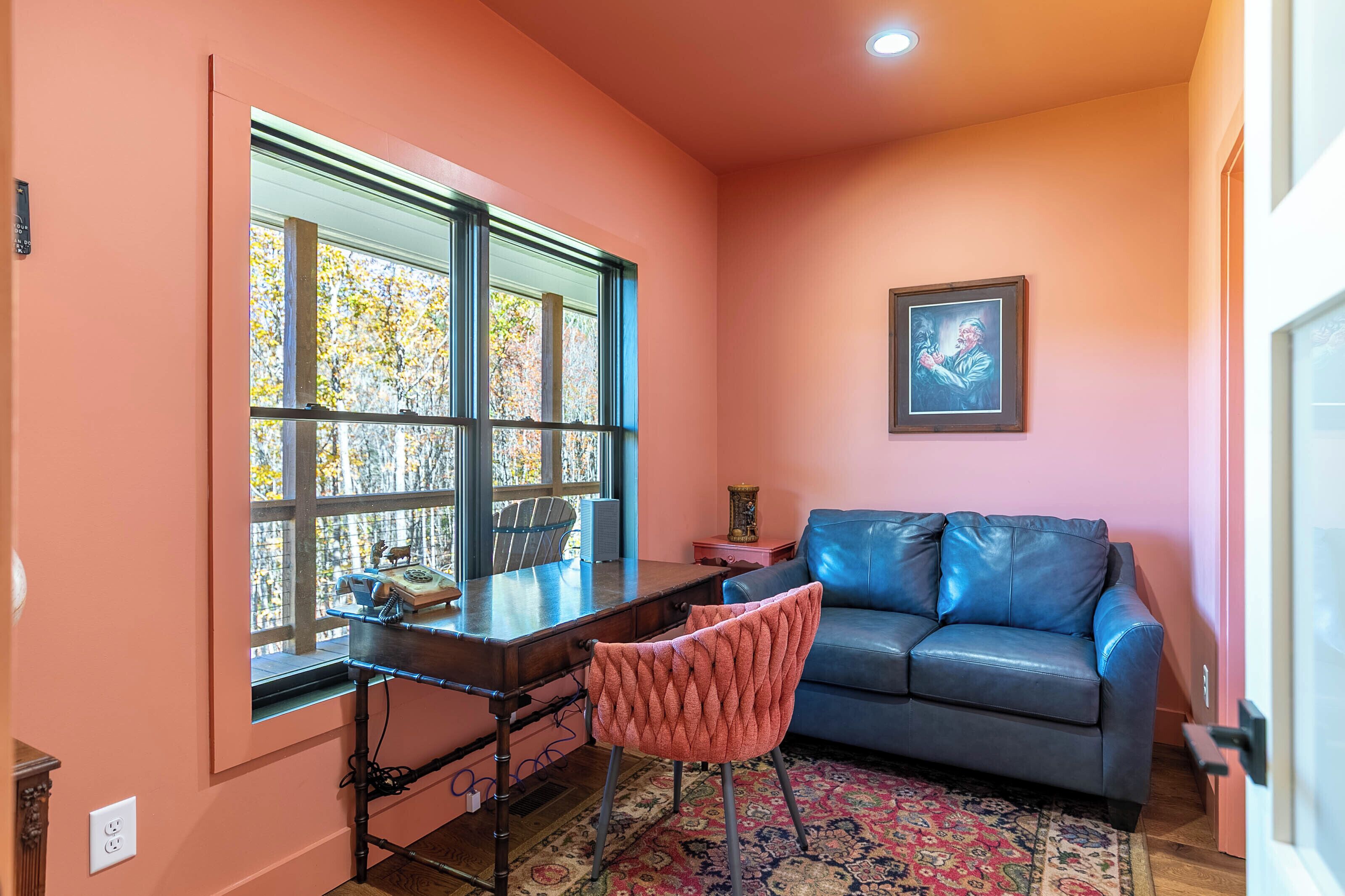 A room with a desk, chair, couch, and a large window with a view of trees.