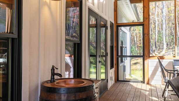 Una terraza de madera con un lavamanos de barril rústico, grandes puertas de vidrio que dan a una vista de bosque, y una zona de comedor con mobiliario exterior.