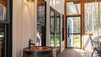 A wooden deck with a rustic barrel sink, large glass doors leading to a forest view, and a dining area with outdoor seating.
