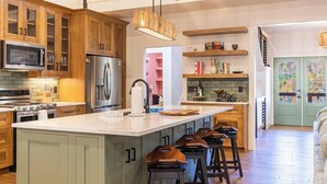 Una cocina con armarios de madera, una isla central, taburetes de bar, un microondas, un refrigerador y una lámpara colgante.