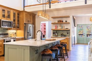 A kitchen with wooden cabinets, a central island, bar stools, a microwave, a refrigerator, and a hanging light fixture.