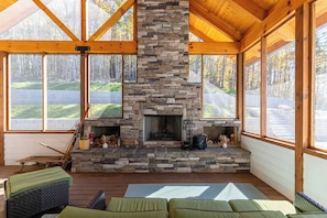 A stone fireplace with a chimney, wooden beams, and large windows overlooking a forested area.