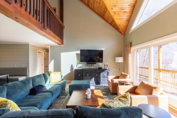 A spacious living room with a large TV, comfortable seating, a wooden ceiling, and large windows. - Mountain View Retreat (Banner Elk)