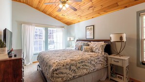 Un dormitorio con techo de madera, cama con ropa de cama floral, mesita de noche con lámpara y ventana con cortinas.