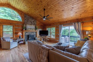 A log cabin interior with a fireplace, wooden walls, a sofa, a television, and a balcony with a view of greenery. - Horse Valley Lodge (Boone)