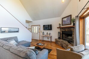 Ein Wohnzimmer mit grauem Sofa, einem Couchtisch, einem Kamin, einem an der Wand montierten Fernseher und einer Treppe, die in einen anderen Raum führt.