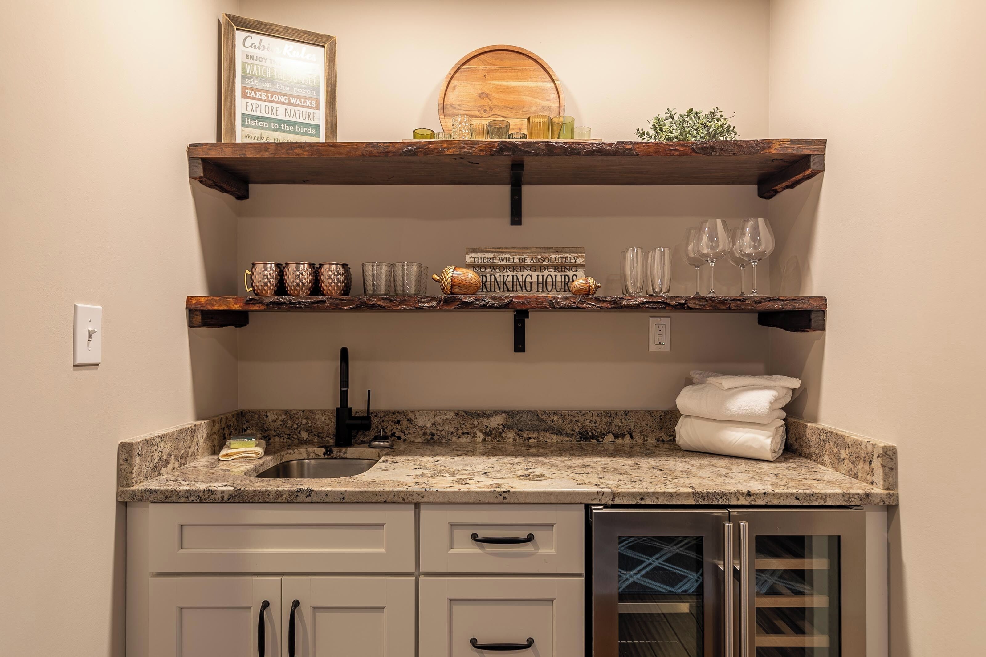 A kitchen with granite countertops, white cabinets, and a stainless steel sink. There are two wooden shelves above the countertop, one holding glassware and the other holding a sign and decorative items.