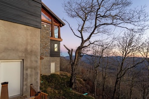Ein modernes Haus mit einer Steinfassade, einer Holzdachterrasse und Blick auf ein bewaldetes Gebirge.
